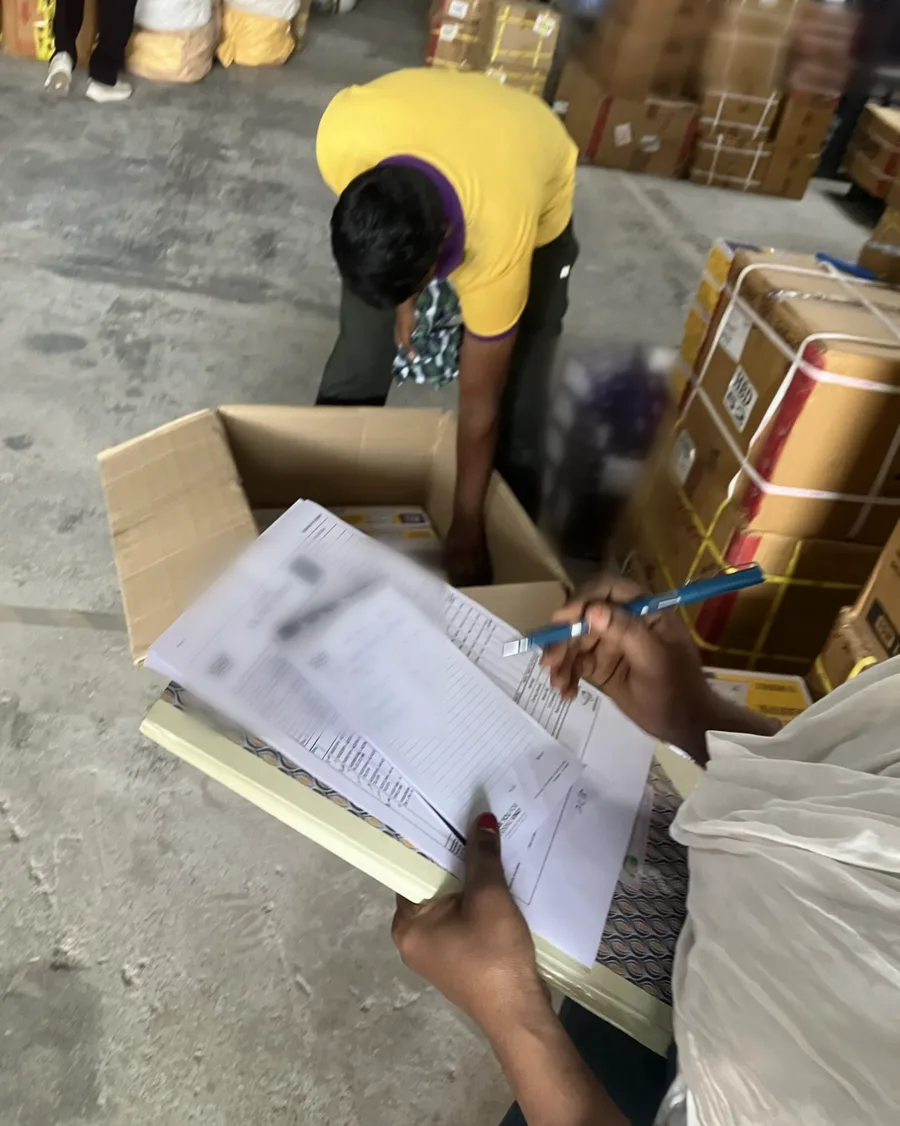 Over-the-shoulder view: a worker picks items into an open carton while someone checks a printed order and handwritten notes on a clipboard; documents blurred.